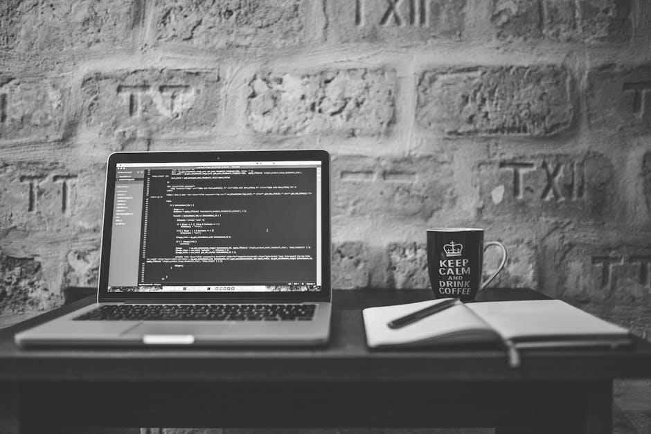 A black-and-white photograph of a workspace featuring an open laptop displaying lines of code on its screen, placed on a dark wooden desk against a textured brick wall background. To the right of the 