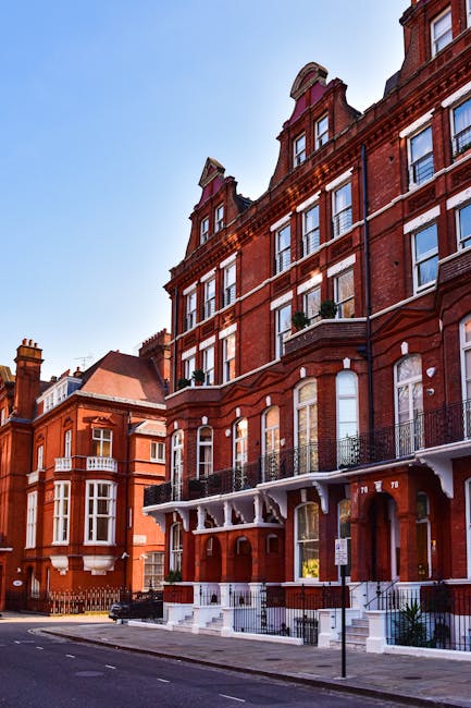 Photograph of a red brick residential building on a city street, showing multiple tall windows with white frames and decorative iron railings on the balconies. The building features ornate architectur