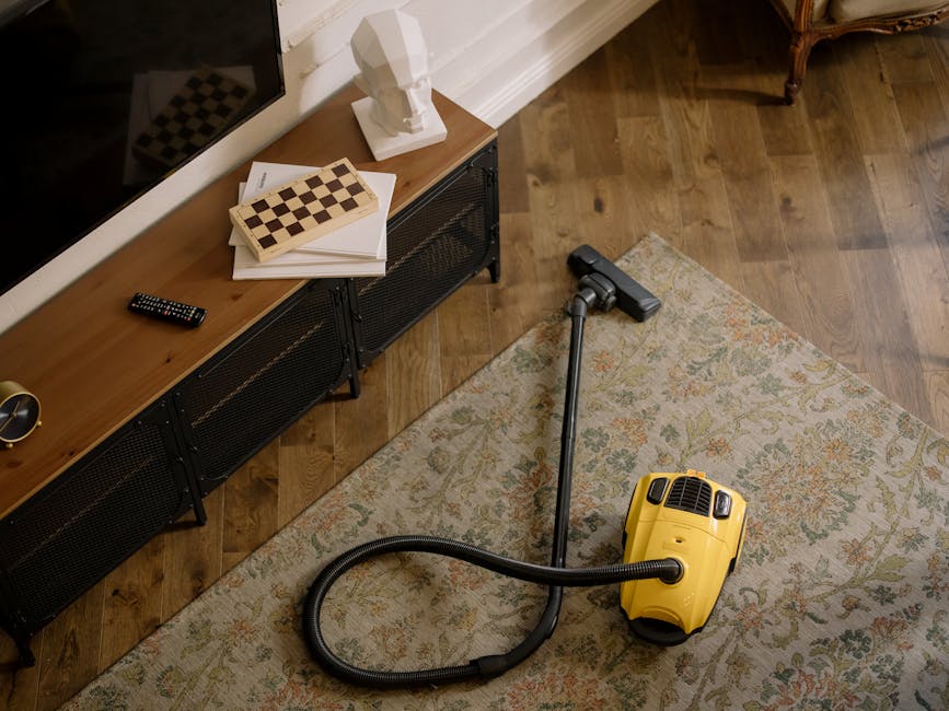 A person using a yellow and black vacuum cleaner with a flexible hose to perform surface cleaning on a patterned carpet in a residential setting. The individual is kneeling on the floor, wearing casua