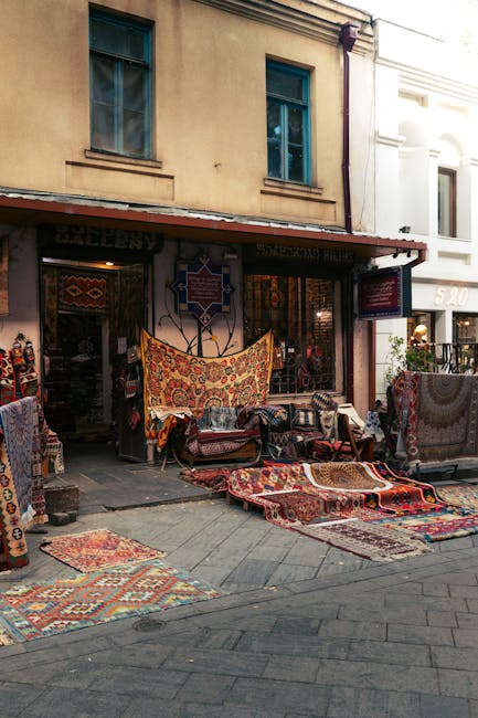 A narrow outdoor courtyard featuring a row of shops or stalls with arched doorways, covered in colorful, patterned textiles hanging from the upper sections. The ground is paved with stone or concrete,